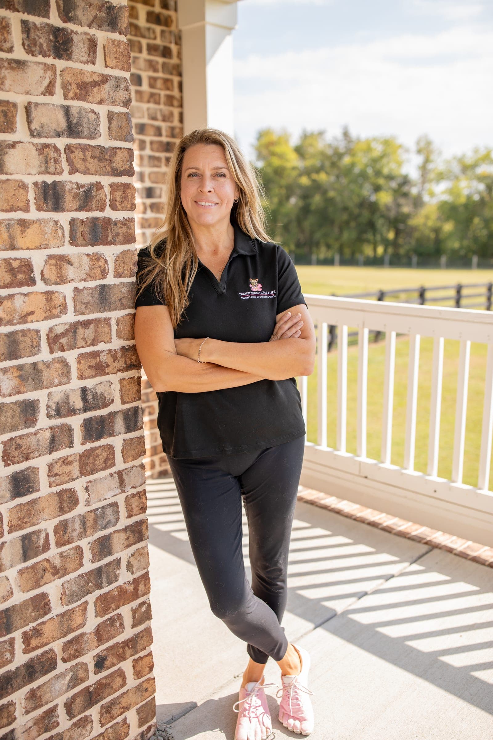 Julie Wilcoxson, nutrition coach in Murfreesboro, TN, standing against a brick pillar on a porch, arms crossed, in a black Transformations 4 Life polo and pink barefoot shoes, a fenced field behind her.